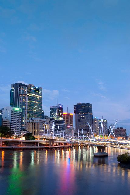 Evening view of the Kurilpa Bridge, Brisbane, a modern pedestrian and cycling bridge over the Brisbane River with the buildings of the CBD on the skyline and colourful lights reflected in the water